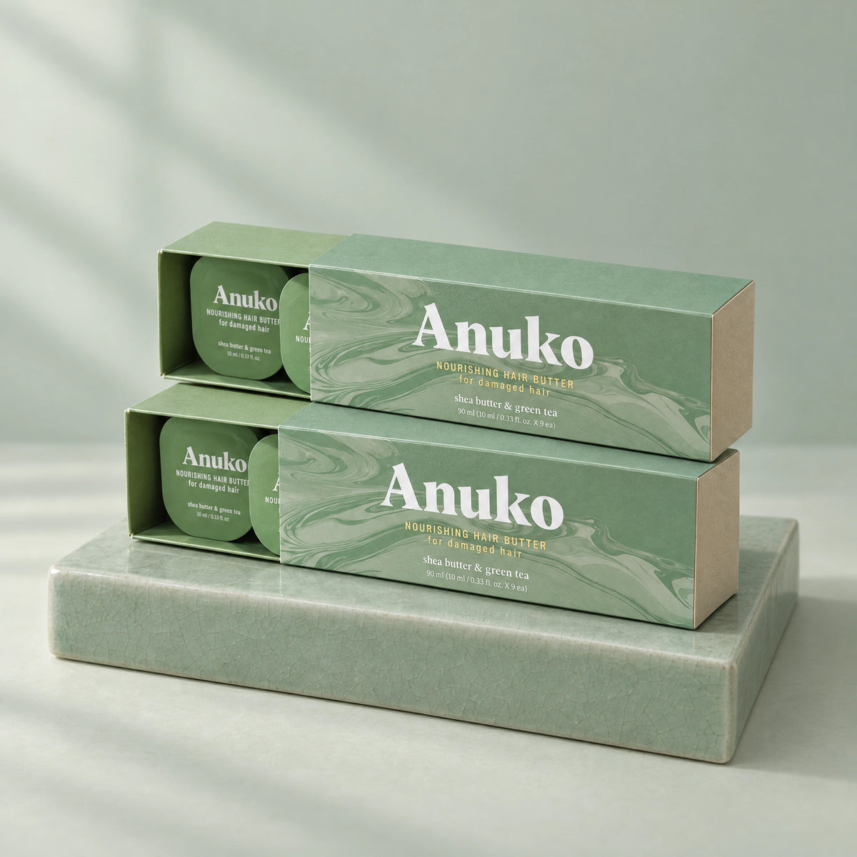 Two Anuko Travel Hair Butter cases stacked on a celadon tile plinth